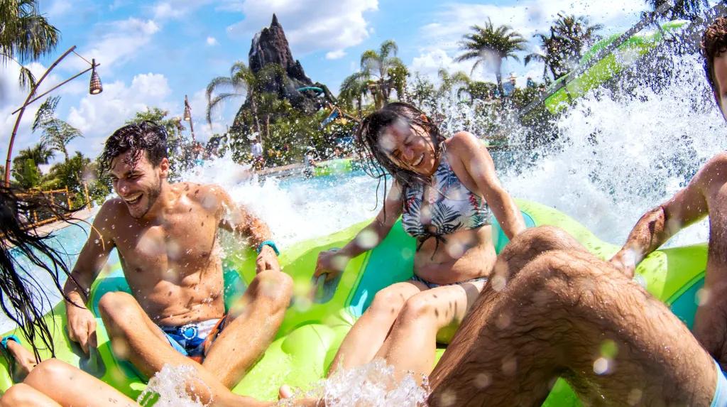Volcano Bay Four young adults sat on a green rubber ring splashing into a large pool with a large statue of a volcano in the background.
