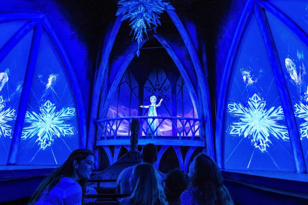 Frozen Ever After Queen Elsa stood on ice balcony surrounded by ice walls and projections of snowflakes. There is a chandelier above hanging made of ice. Below there are guests sat in rows on boat.