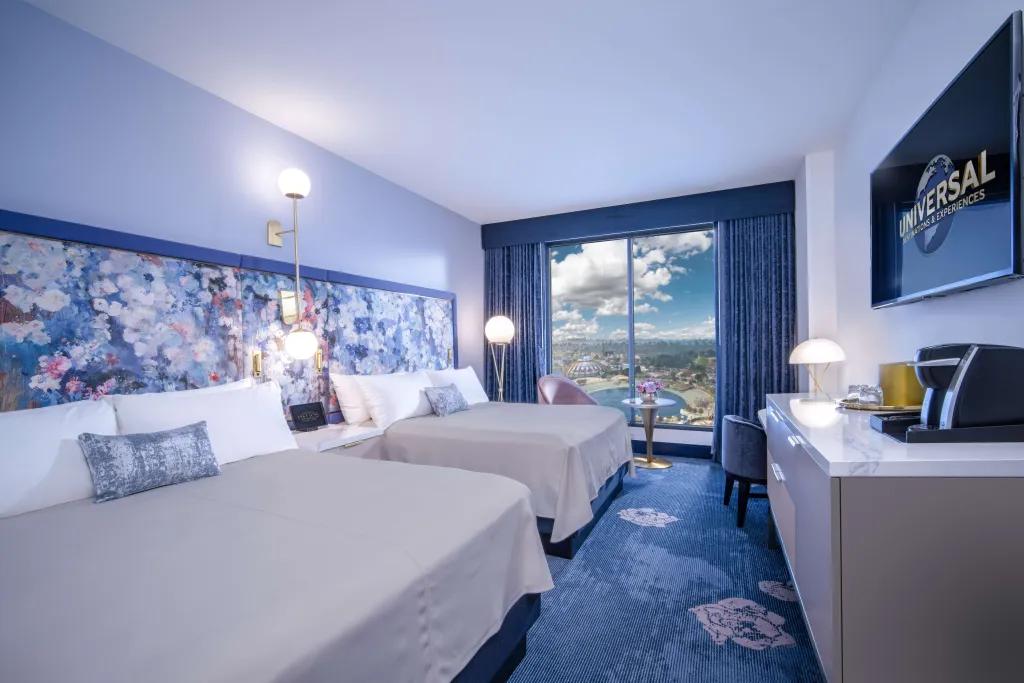 Universal Helios Grand Hotel Room Blue room with two double white beds side by side in spacious area. There is a tv on the wall with the Universal sign on and there is a big window with view of Epic Universe. On the wall there is a floral pattern in a blue hue.