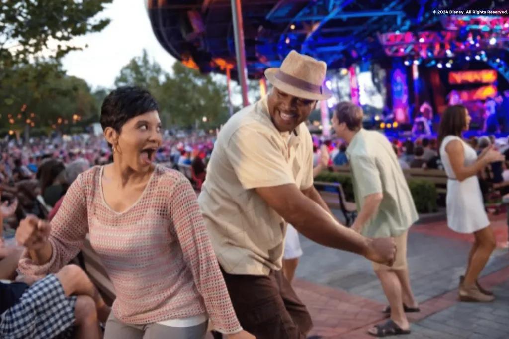Eat To The Beat Woman laughing dancing back to back with a man with other people dancing in the background. There is a stage as well.