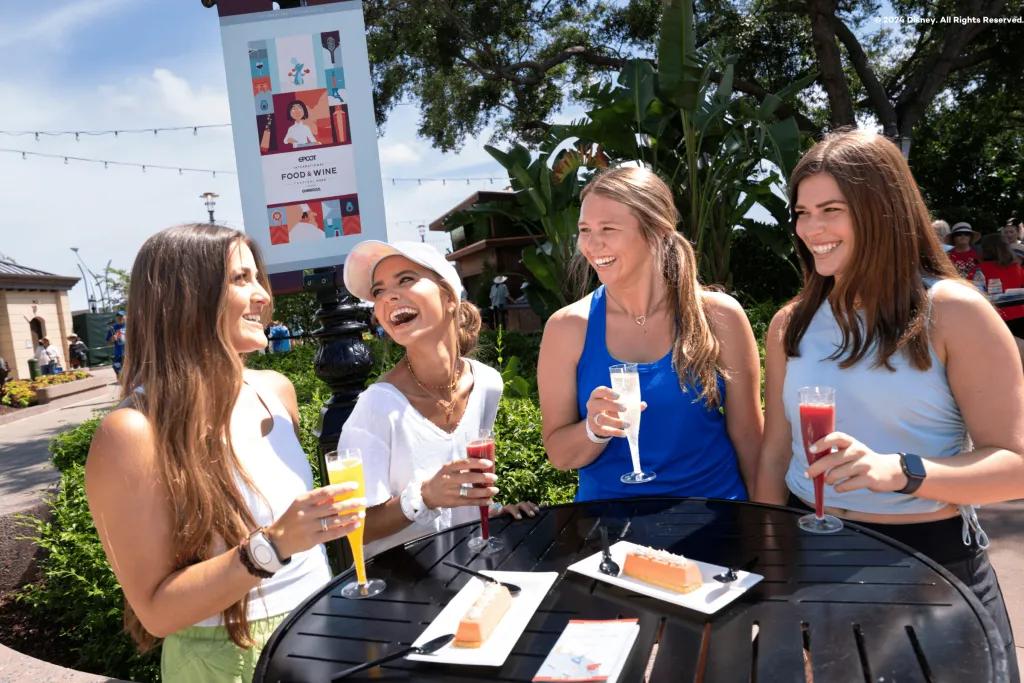 EPCOT Food & Wine Festival Four girls looking at one another laughing as they are sat a table holding different coloured plastic glasses of drinks. Two are red, one yellow and one white. There are plates of perfect looking desserts on the table with plastic knives and forks.