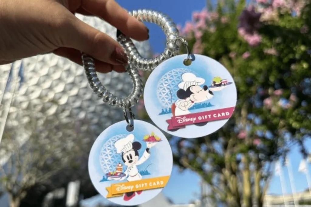 Disney Gift Card wristlet Two see through wristbands held up by a hand, on each are designs of blue backgrounds and one has Mickey Mouse and the other has Minnie Mouse. Both characters are wearing chef costumes and holding meals. Text on it says Disney Gift Card. Spaceship Earth is in the background.