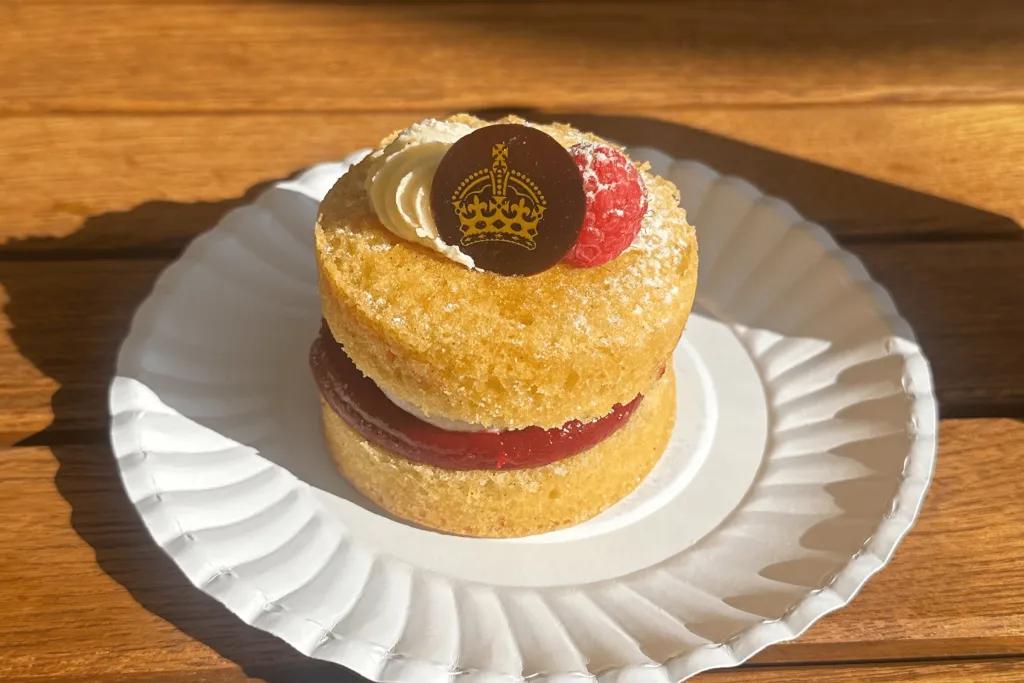 Victoria sponge cake in Buckingham Palace Small, circular Victoria sponge cake served on paper plate. It is two vanilla cake slices with cream and jam in the middle. On top there is cream, a raspberry and a circular piece of chocolate with golden image of crown on it.