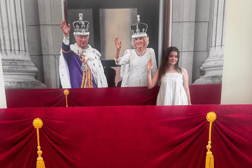 Photo opportunities in Buckingham Palace Girl stood in front of image of King Charles and Queen Camilla stood on the balcony during coronation with crowns on their heads. They are both waving and so is the girl waving and smiling. There is also a cardboard red velvet curtain in front to replicate balcony.