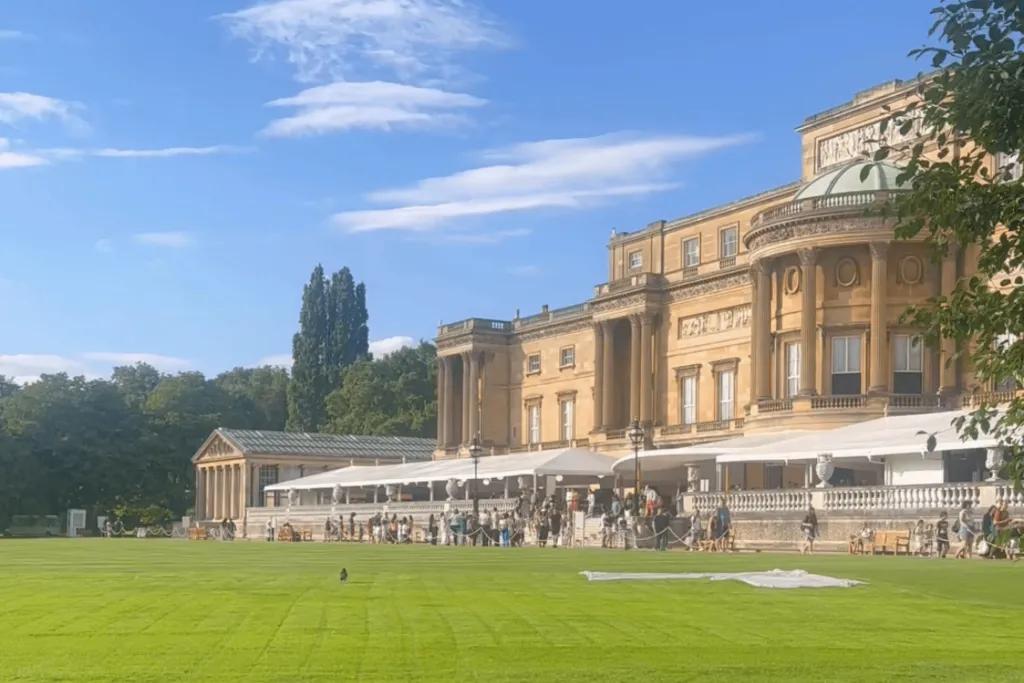 Buckingham Palace Gardens The back of Buckingham Palace, a large beige building which backs onto large green grass patch and trees. There is also a white tent with cafe in.