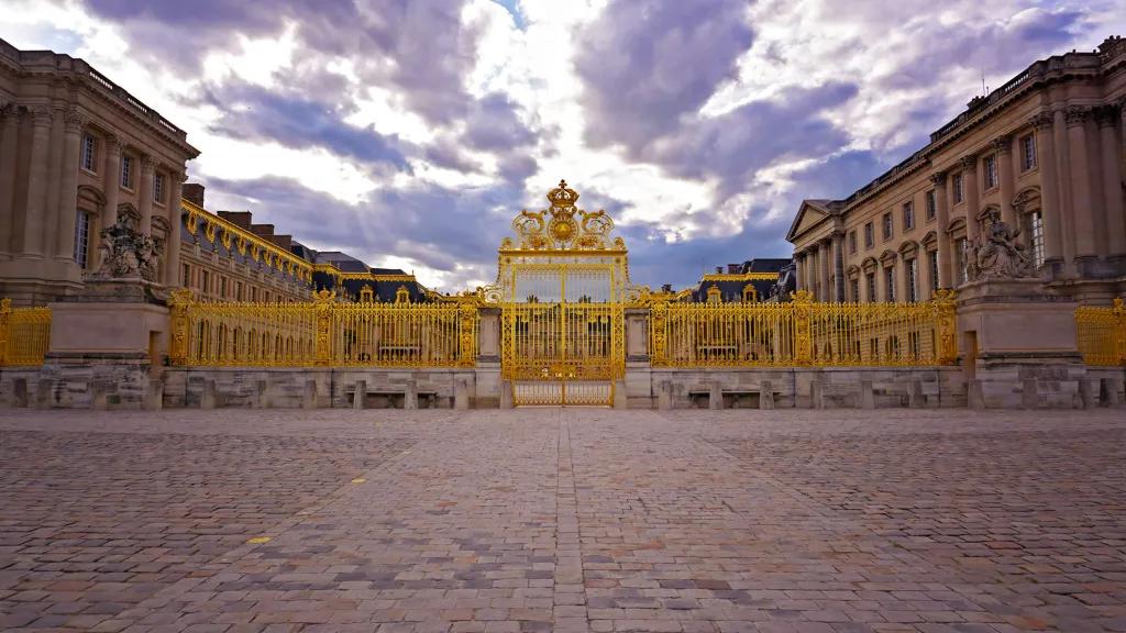 Palace of Versailles Golden gates with beautiful royal details, there are stone statues and behind is a grand palace.
