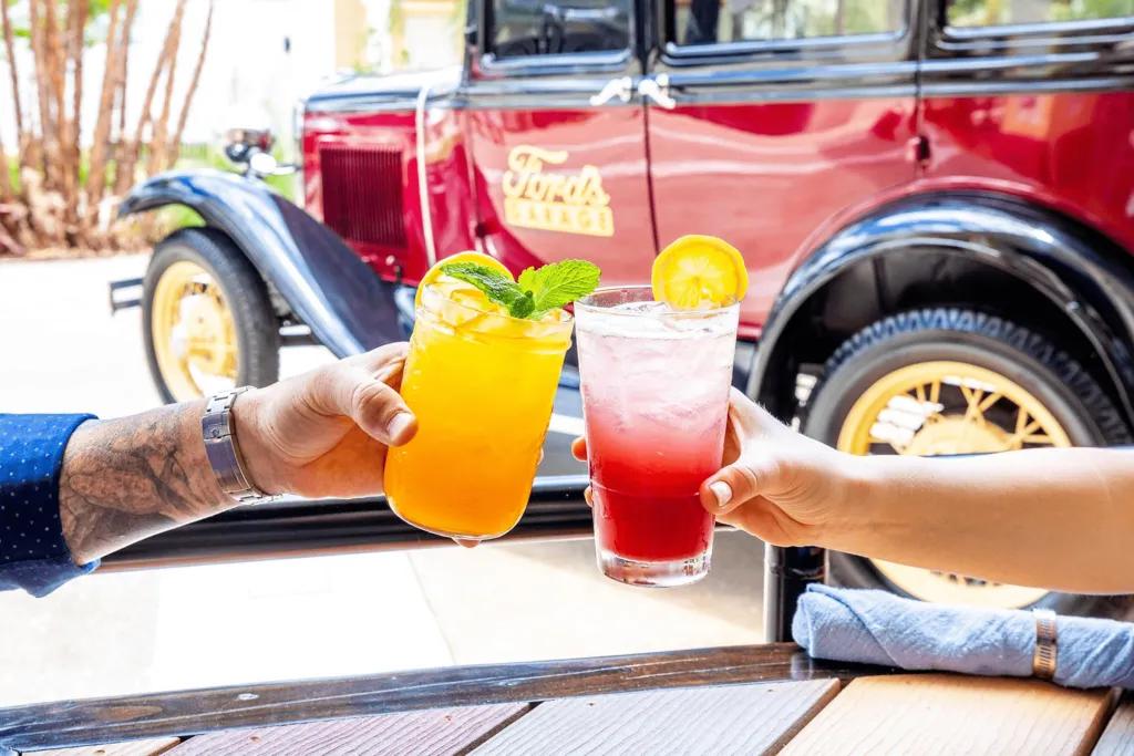 Ford's Garage Two people clinking glasses of cocktails in front of an old fashioned car with Ford's Garage on the side
