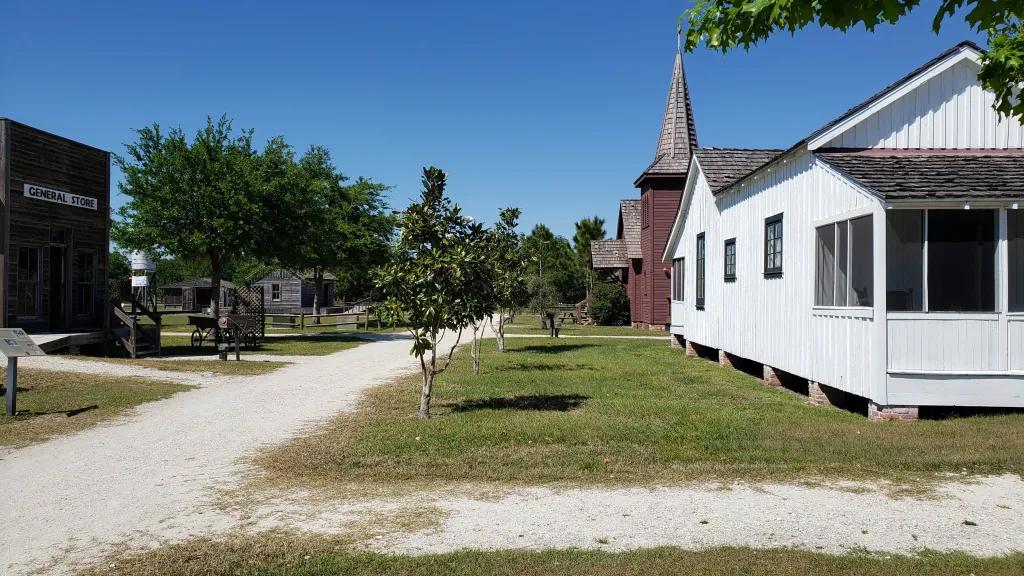 Pioneer village Kissimmee Deserted street with a white house on the corner and behind there is a grey building which has a pointy roof, looks like a church. There are small trees on the side of dirt track road.