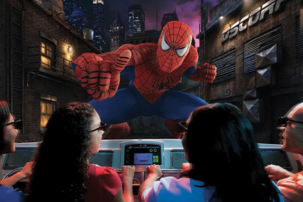 The Amazing Adventures of Spider-Man 4 people sat on ride vehicle. They are wearing black 4D glasses and it looks like a man stood on the front of the vehicle in spiderman suit. The suit is red with black spider webs on and blue legs.