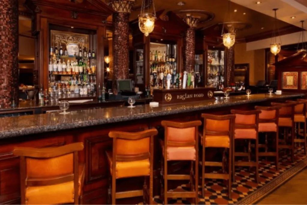 Raglan Road Dark wooden bar with orange and wooden bar stools. There are drinks hanging behind the bar and it is completely empty.
