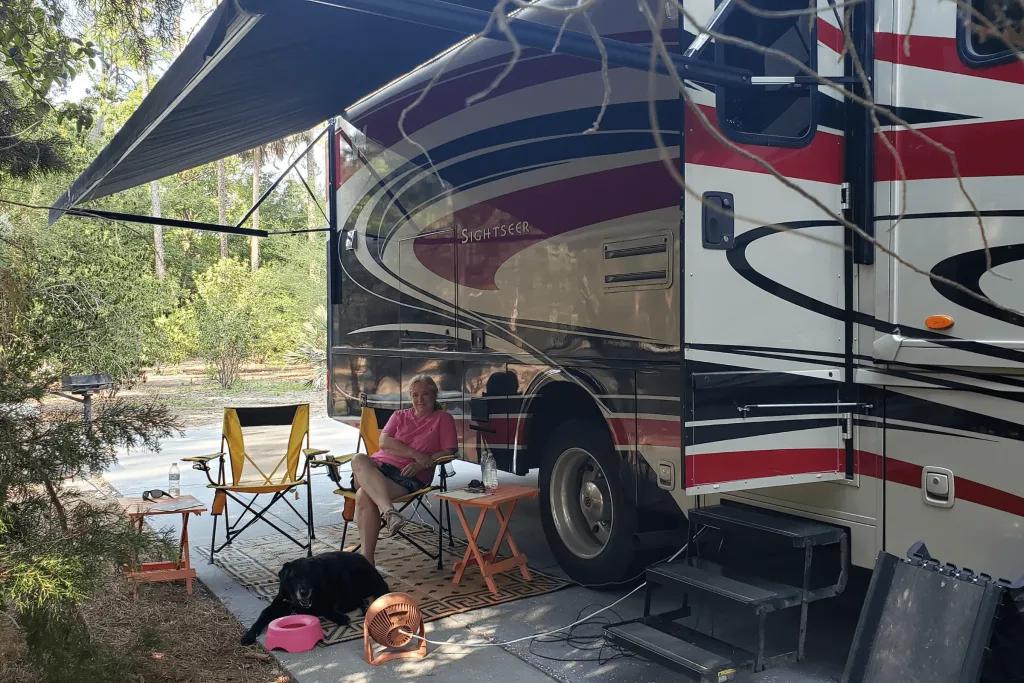 Disney's Fort Wilderness Woman sat on foldable chairs outside a campervan under some shade. The door is open and the black dog is sitting at the woman's feet.