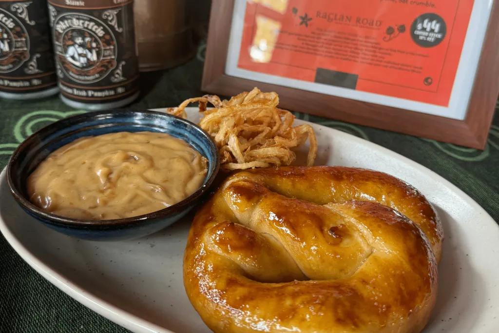Food at Oktoberfest Potato Pretzel with a yellow mustard looking sauce and crispy food on the side. There are Oktoberfest cans in the background with beer in them.