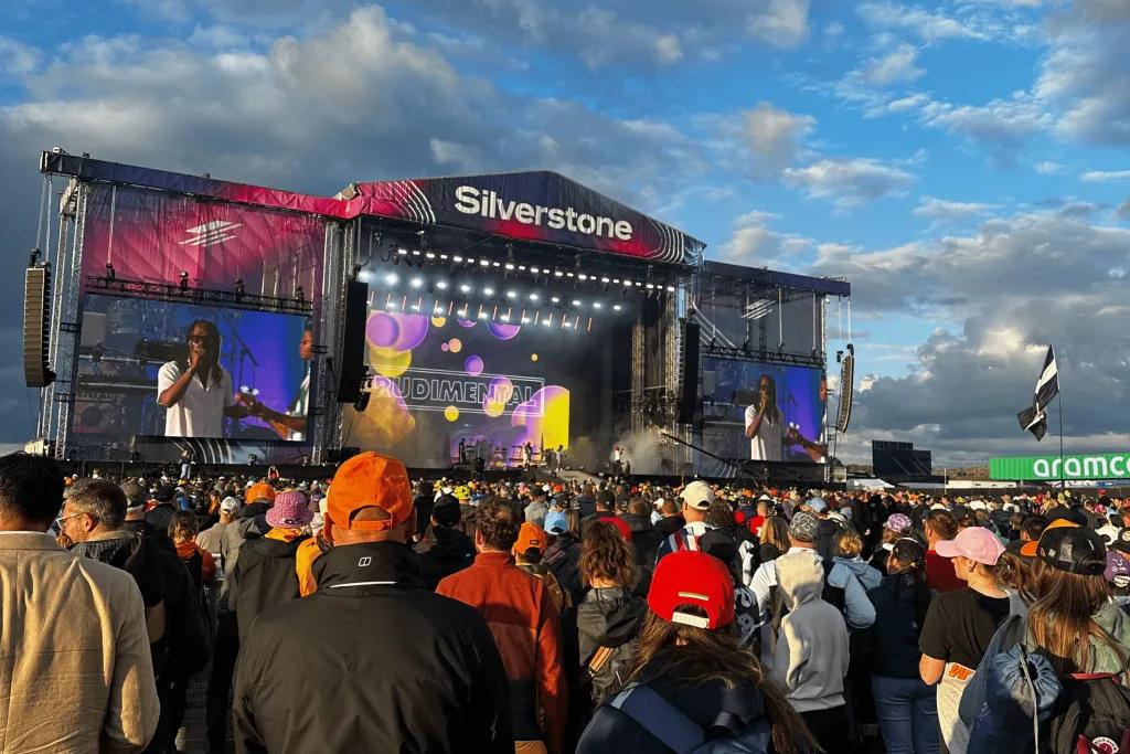 Silverstone Grand Prix There is a large stage with multi-coloured lighting on it. There is a large crowd watching the stage wearing different F1 teams merch. The stage says Silverstone at the top of it.