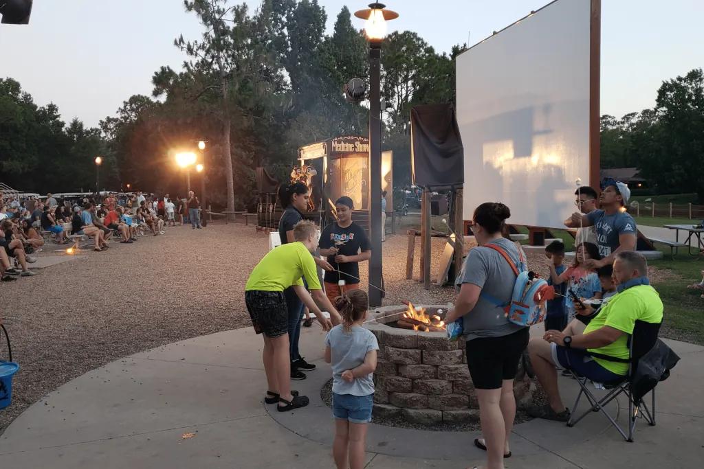 Campfire Sing-Along with Chip ‘n’ Dale Guests are gathered around a campfire roasting marshmallows on sticks. There are many tables full of guests and the sun is setting over the camp site. There is a little stage for Chip 'n' Dale to perform on and a board for movies to be projected onto.