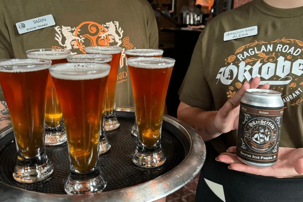Oktoberfest Two workers holding drinks and wearing dark green t-shirts which say Oktoberfest and have nametags on. One is holding a black tray with large shots on and the other is holding a silver can which has Oktoberfest theming on it.