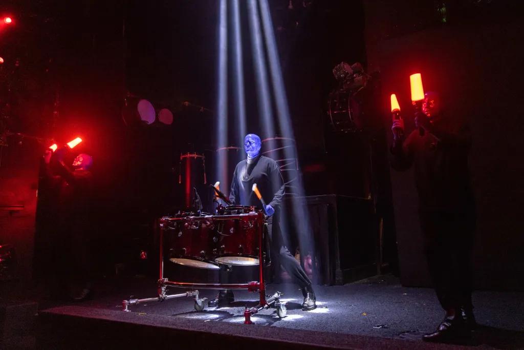 Blue Man performing There is a man dressed in all black and painted completely blue holding two sticks and has a wooden instrument in front of him. There are 4 small spotlights shining down on him centre stage.