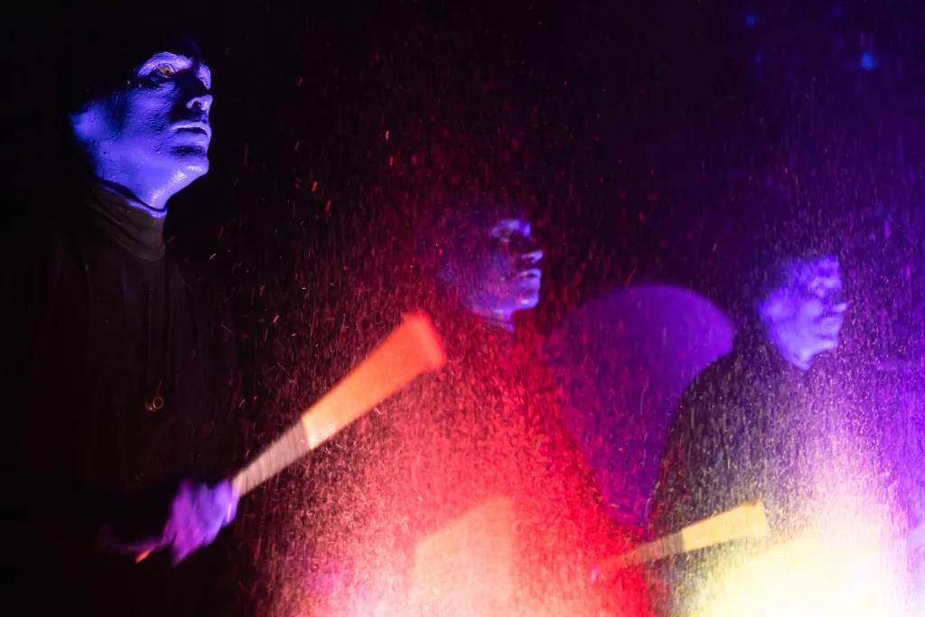 Blue Men Show Three men painted completely blue wearing black outfits are holding glowing sticks and smacking a drum which has loads of neon paint of different colours.