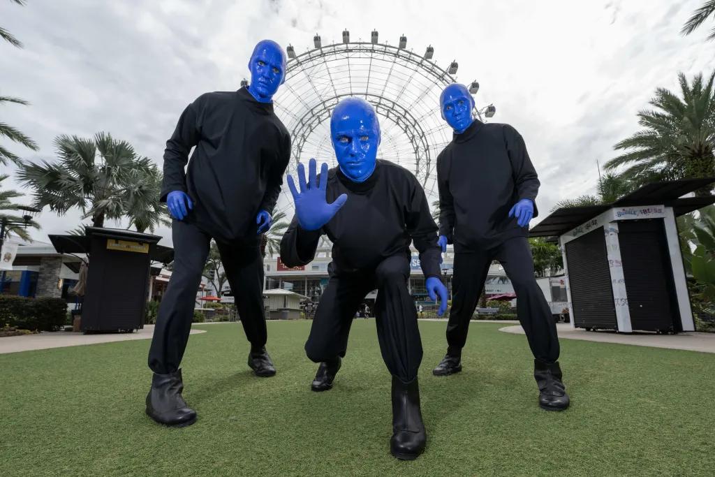Blue Men Group at Icon Park There are three men completely painted in blue paint stood in front of a big white Ferris wheel on a field. Each of them are posing in a strange way.