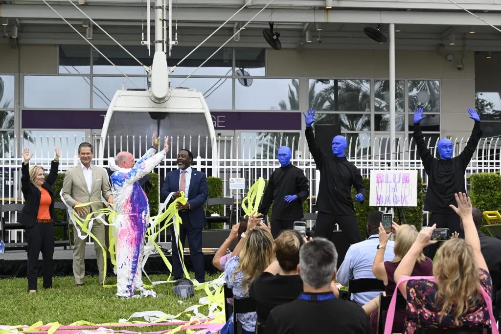 Blue Man Group Press Conference Three men dressed in all black and painted completely bright blue stood with their arms up looking at a man in an all white suit with paint splattered all over him. Behind there are Ferris wheel carriages at the station and there are many people sat in rows of seats cheering along.