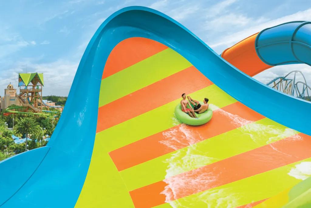 KareKare Curl Two men sat on two seater lime green rubber ring on a blue slide with lime green and orange stripes. They are heading up a large curled up wall.