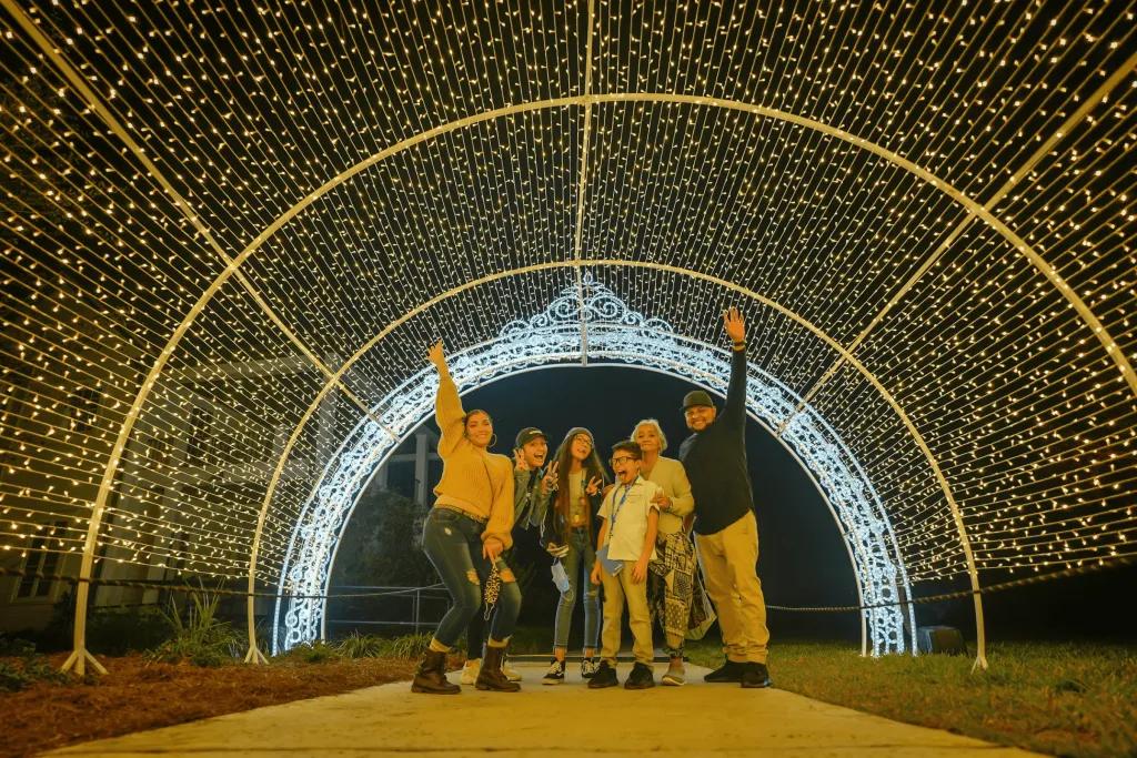 Leu Gardens Family of 6 stood under a tunnel of lights at night. They have their arms lifted up towards the magical yellow and white lights.