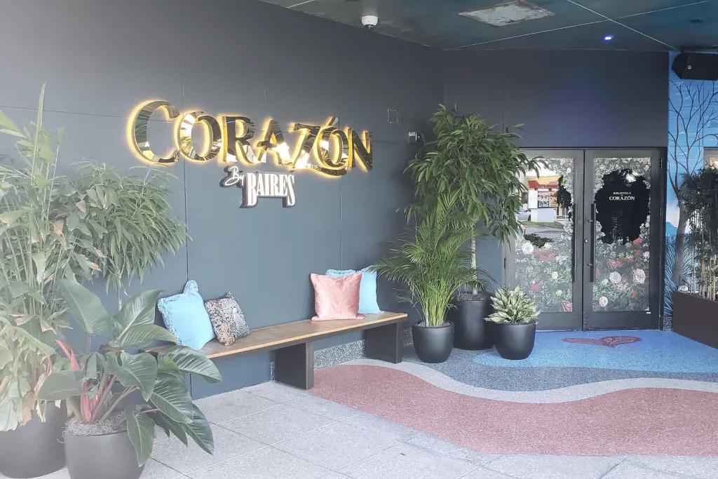 Corazón by Baires The entrance to the restaurant with a golden sign saying corazon lit up on the dark grey wall underneath there is a wooden bench with pillows. To the right there are window door.