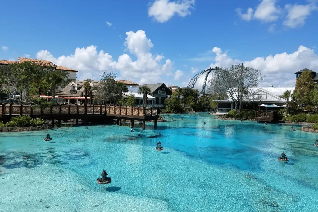 Disney Springs There is a crystal clear lake with fountain cannons. In the background there is a wooden pier and a spherical metal building.