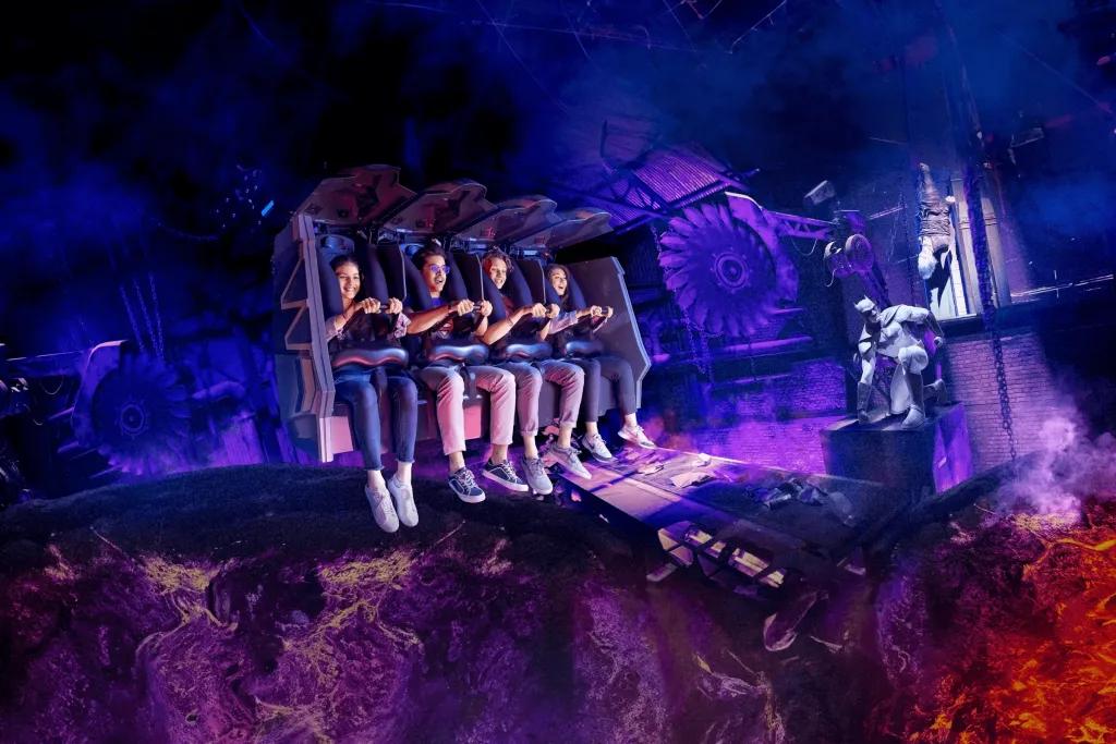 Batman: Knight Flight Group of four sat on rollercoaster car held in by lap bars with their legs hanging. In the background you can see Batman sat in a dark and dingy area.