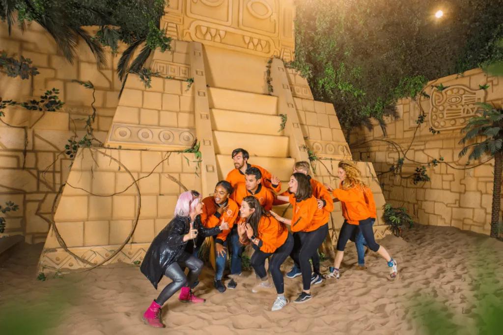 Crystal Maze Woman wearing wacky outfit and pink wig is crouching with a group of guests who are wearing matching orange jumpers. They are stood on sand are in front of two large temple.
