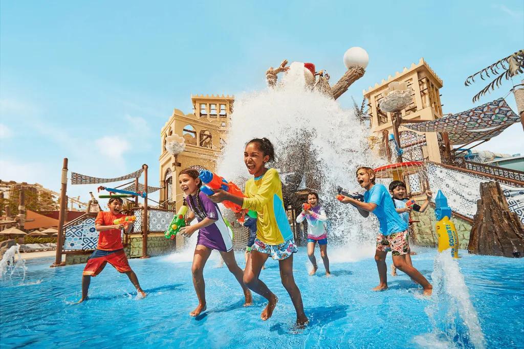 Marah Fortress There are many little kids running away from play area large bucket pouring huge wave of water down onto them. They are all holding water guns and laughing pointing them at one another. The play area has a large climbing frame in a shallow pool.