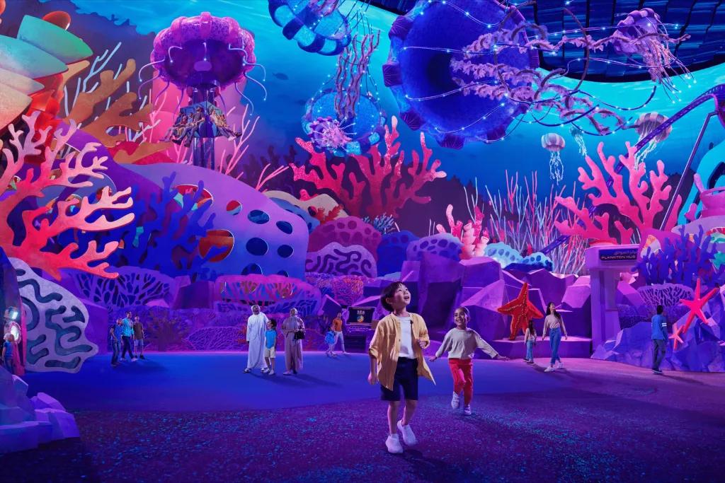 SeaWorld Abu Dhabi Young boy walking through a large room with luminous decorations in it. There's large coral reef decorations and flying overhead are hanging sea weed decorations.