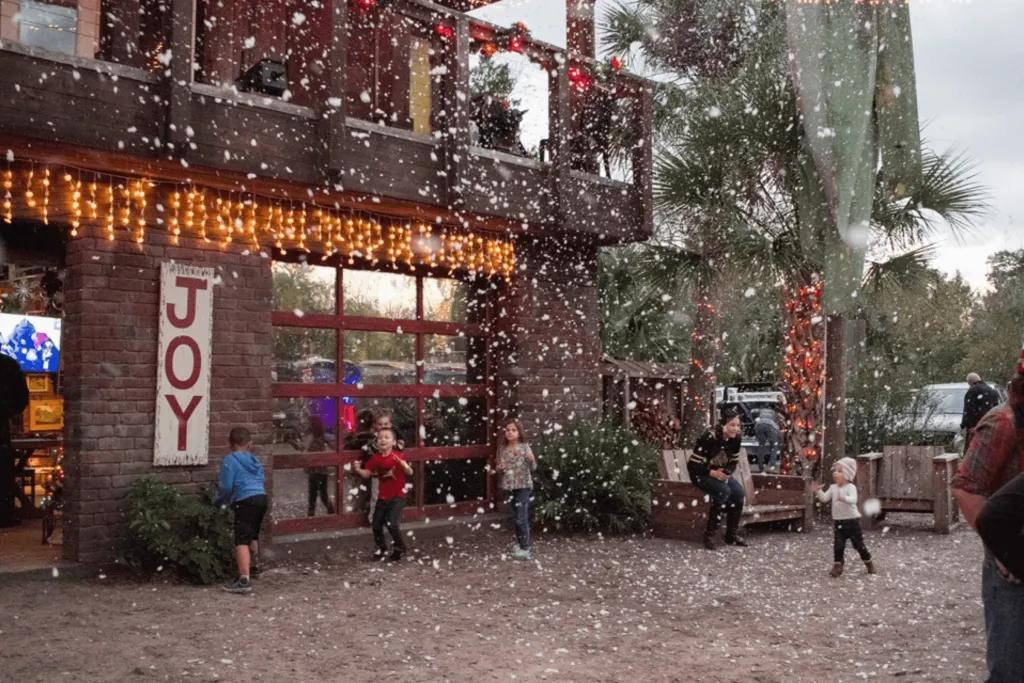 Wekiva Island Christmas Stone building with Christmas decorations including orange lights and a sign saying JOY on it. There is fake snow floating down and young kids playing in it.