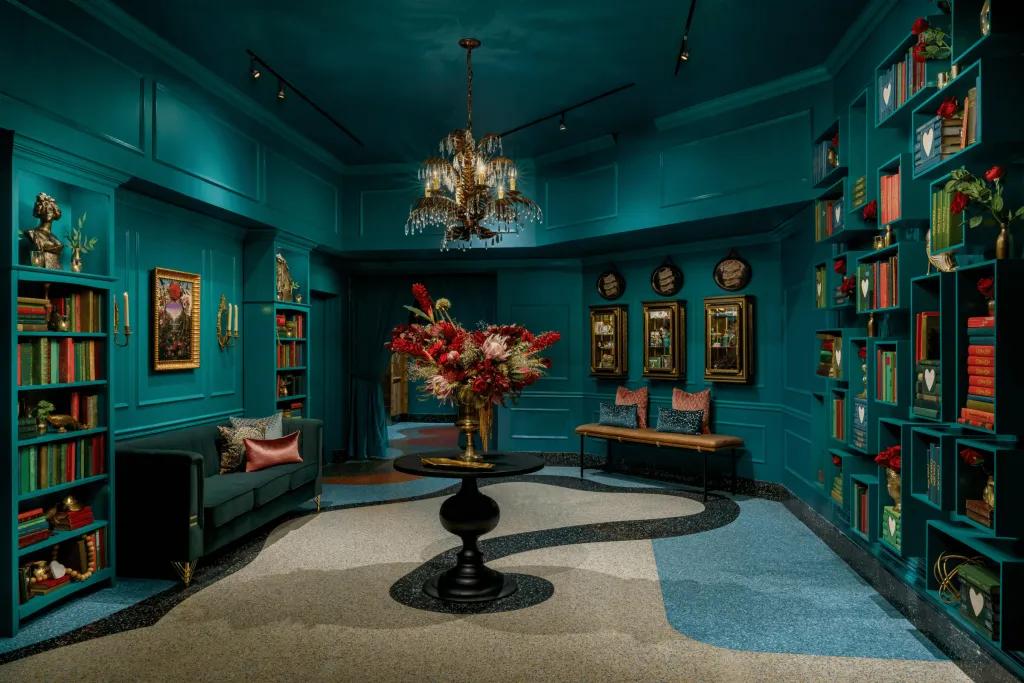 Corazón by Baires, image by Dylan Cross Turquoise large room with many shelves full of books on the wall, an obscure white, blue and black swirling pattern. In the centre there are flowers in a large vase.
