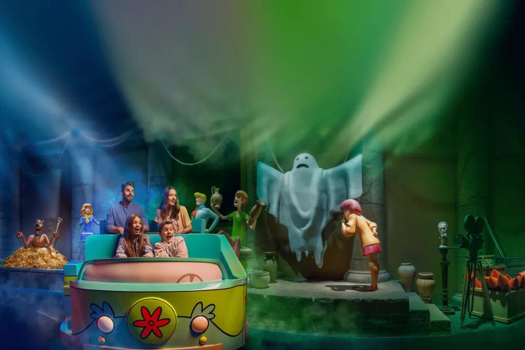 Scooby Doo: The Museum of Mysteries A family of four are sat in a ride vehicle with two rows of seats painted like the car from Scooby Doo. In the background there is the gang from Scooby Doo and Velma is whispering in the ear of a ghost with its arms held in the air.