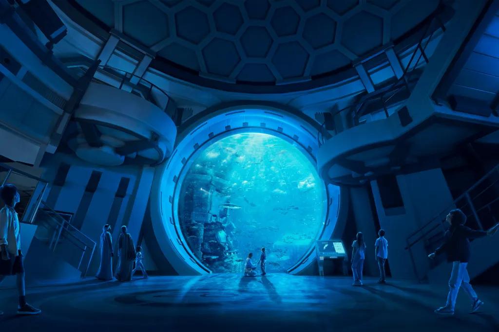 Aquarium in SeaWorld Abu Dhabi Mum kneeling and holding hand of child stood in front of a huge circular window. On the other side there are fish and marine animals swimming around. The room is dark and there are a lot of guests walking around.