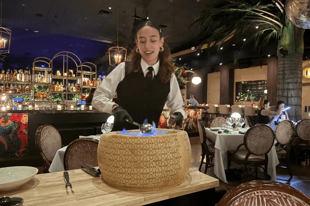 Corazón by Baires, image by Matt Roseboom Waitress dressed in black and white suit and waistcoat dishing food in a giant wheel cheese and holding a large spoon which is lit on fire. In the background, you can see the classy decor of the restaurant with the bar and palm trees lit with sparkling lights.
