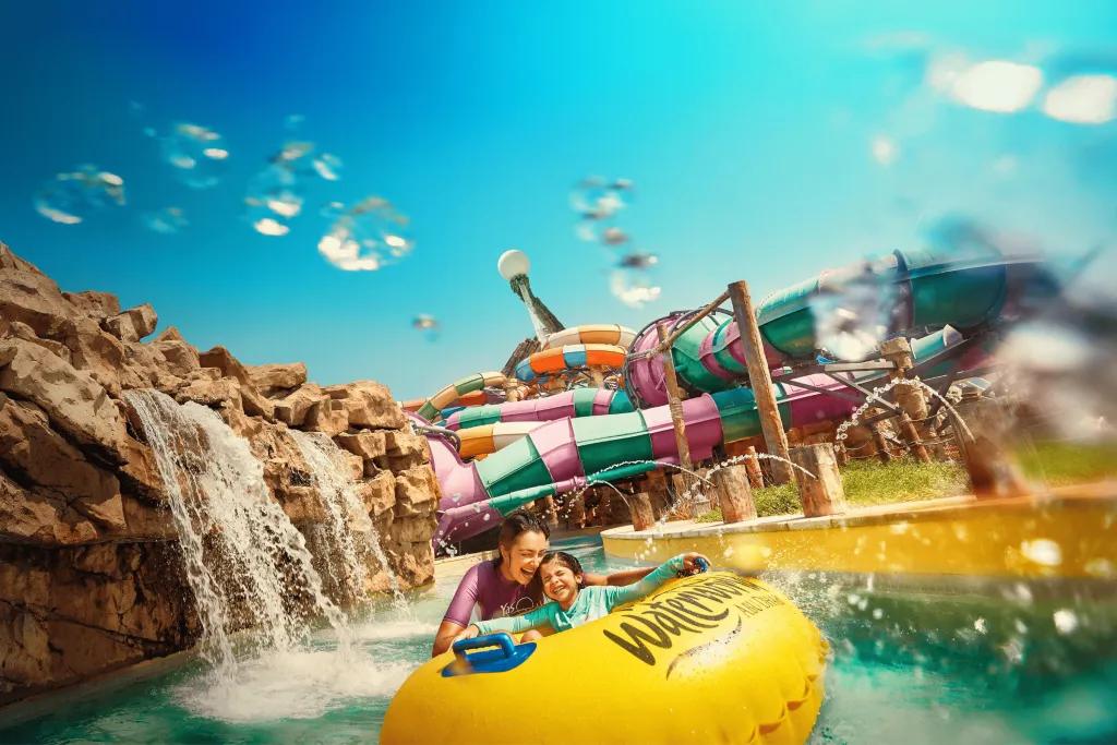 Yas Waterworld In the background there are many large waterslides wrapping around one another in different colours. There is a small lazy river flowing with a waterfall pouring into it off of rocks. There is a mother holding her little girl who is sat on a yellow rubber ring. There are fountains also splashing them.