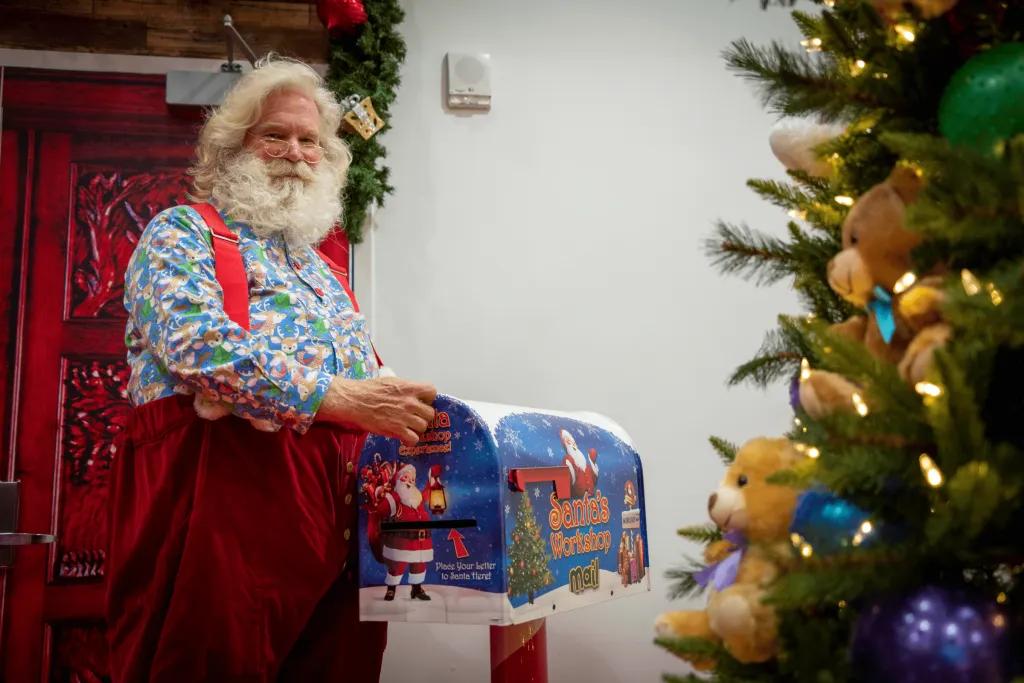 ICON Park Santa Workshop Santa, a man with blue patterned shirt on and red dungarees on over the top made of velvet. He has white hair and a white beard. He is holding a gift and putting it under the Christmas tree, decorated in gold and blue decorations.