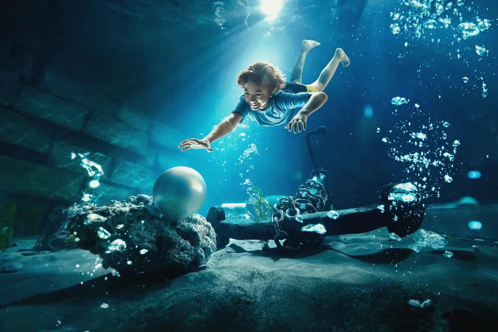 Hairat Yas Pearl Diving There is a young woman diving down into a deep pool reaching out to a large white pearl sat at the bottom of the pool. There is also an anchor sat at the bottom of the pool and bubbles surrounding the woman.