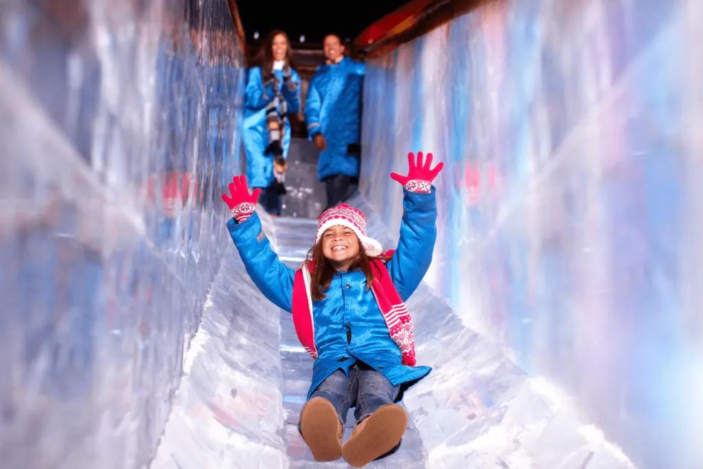 Gaylord Palms ICE! Slide Slide made of ice with a young girl wearing a long blue jacket, red santa hat and red gloves. She is smiling with her arms lifted in the air.