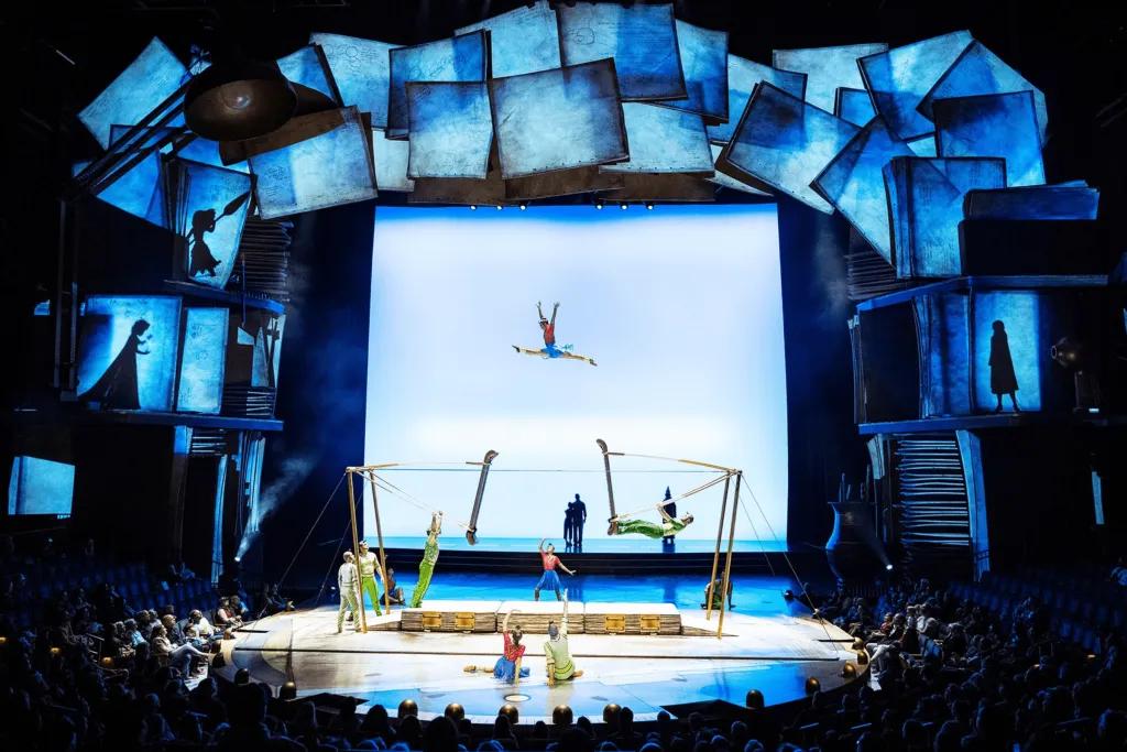 Drawn to Life A large stage lit in blue lights and incredible theming with pages surrounding it. There are gymnasts on the stage with one leaping in the air in the splits.