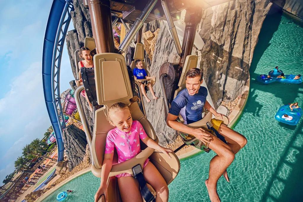 Bandit Bomber A child and her dad sat on a hanging coaster in their swimwear smiling and laughing as they glide above the large swimming pool. Below there are people lounging on rubber rings in the lazy river.