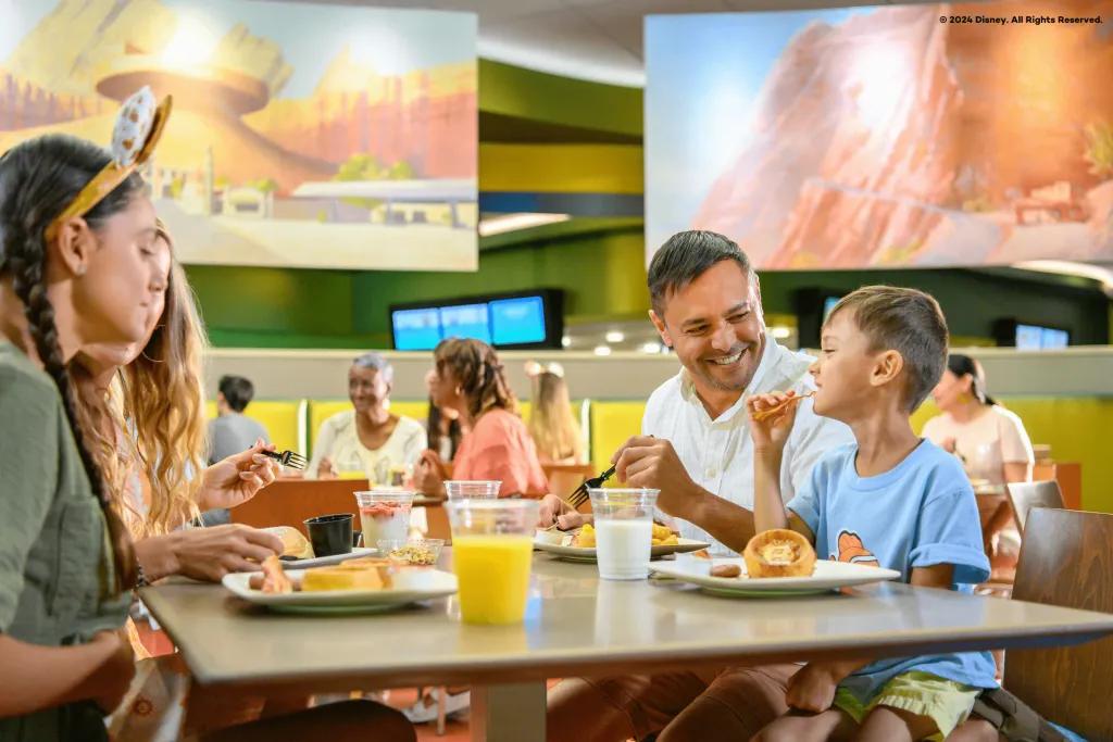 Disney's Art of Animation Resort Dining Plan Family eating quick serve breakfast together in Disney's Art of Animation Resort. Little boy is eating bacon with his hands and the dad is smiling at him.
