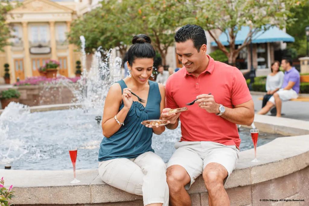EPCOT Festival Dining Couple dining sat on the stone edge of the fountain in the French Pavilion in the world showcase. They have small plates of food and glasses of sparkling red drinks. They are both smiling.