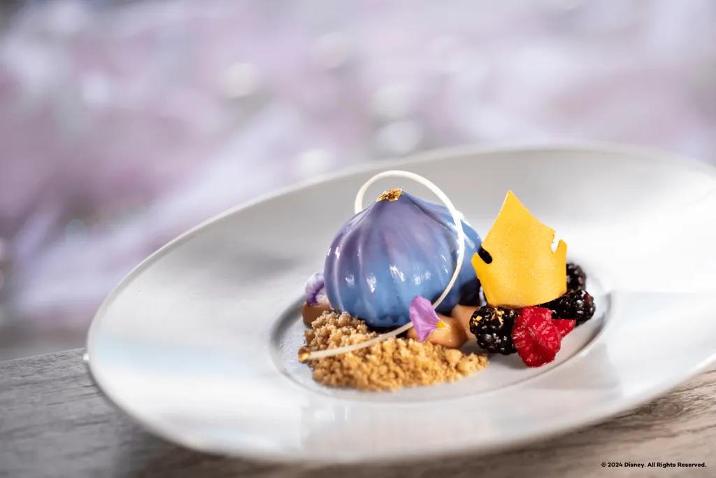 Topolino's Terrace in Disney's Riviera Resort Blue and purple droplet shaped dessert with gold on top. There is also biscuit crumbs at the bottom, fruit beside and a golden crown on top.