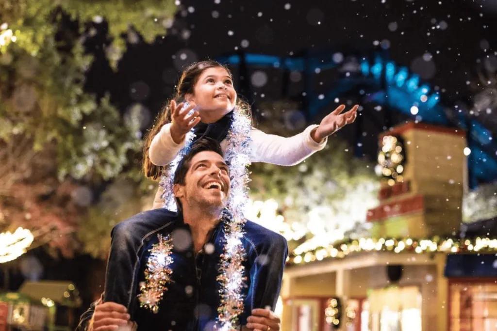 SeaWorld Orlando Christmas Dad looking up at his daughter who is sat on his shoulders and wearing tinsel around her neck. The little girl is reaching out to touch the fake snow. Behind there are trees and buildings decorated with lights and reefs.