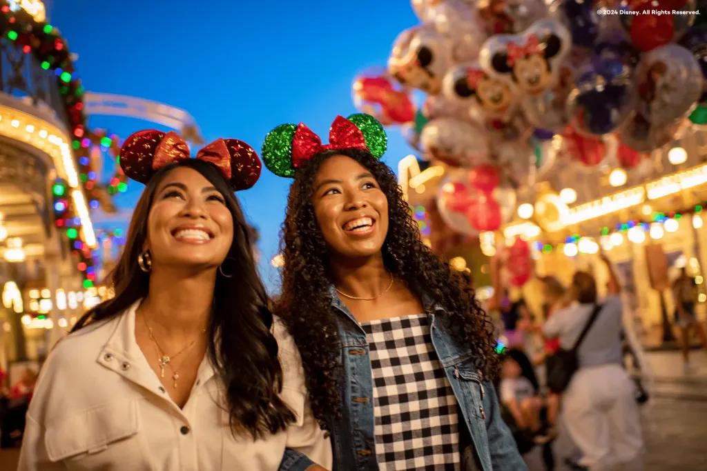 Mickey's Very Merry Christmas Party Two young women with sparkly green and red mickey ear headbands on stood on main street. The buildings in the background are lit and there are balloons in the background.