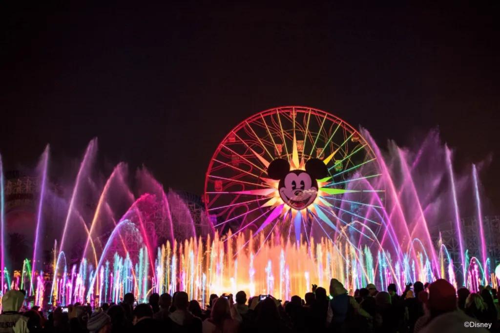 Disney California Adventure Park Huge ferris wheel lit up in the different colours of the rainbow with huge Mickey Mouse's face smiling in the middle. In front there are huge fountains which look like they're almost as tall as the Ferris wheel lit up pink. There are so many.