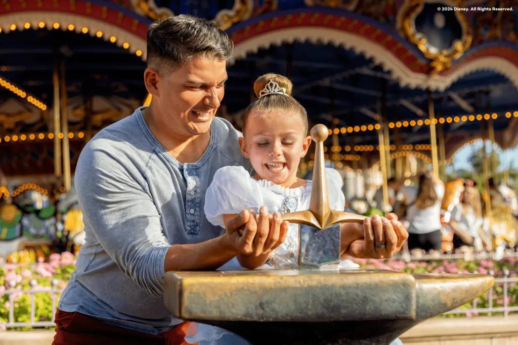 The Sword in the Stone Dad helping his daughter who is dressed in a blue princess dress, pull out a sword from a stone. They are stood in front of the carousel.