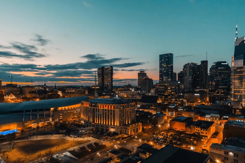 Nashville A city at sunset with all of its lights o. There are many sky scrapers and a few smaller buildings. The lights are an amber colour matching the sky.